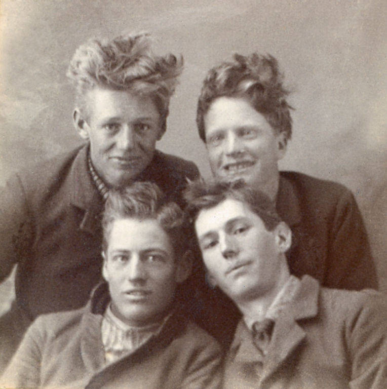 Four young men are closely gathered together, looking at the viewer. They are wearing jackets and shirts, and all have tousled hair. The overall mood appears casual and friendly.