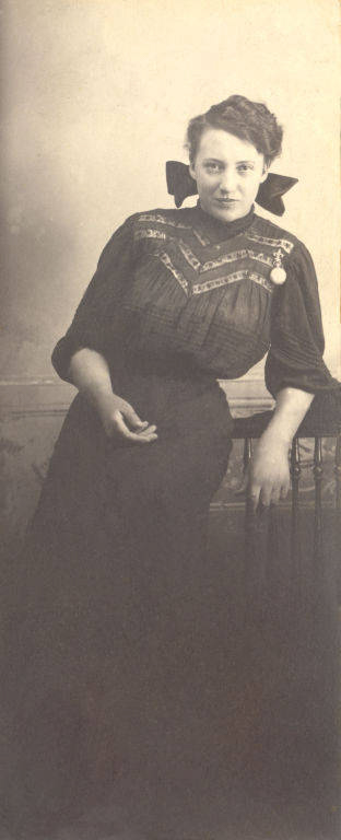 A woman leaning against a railing, wearing a dark dress with decorative patterns on the upper part and a large bow at the neck. Her hair is styled up, and she appears relaxed. The background is plain.