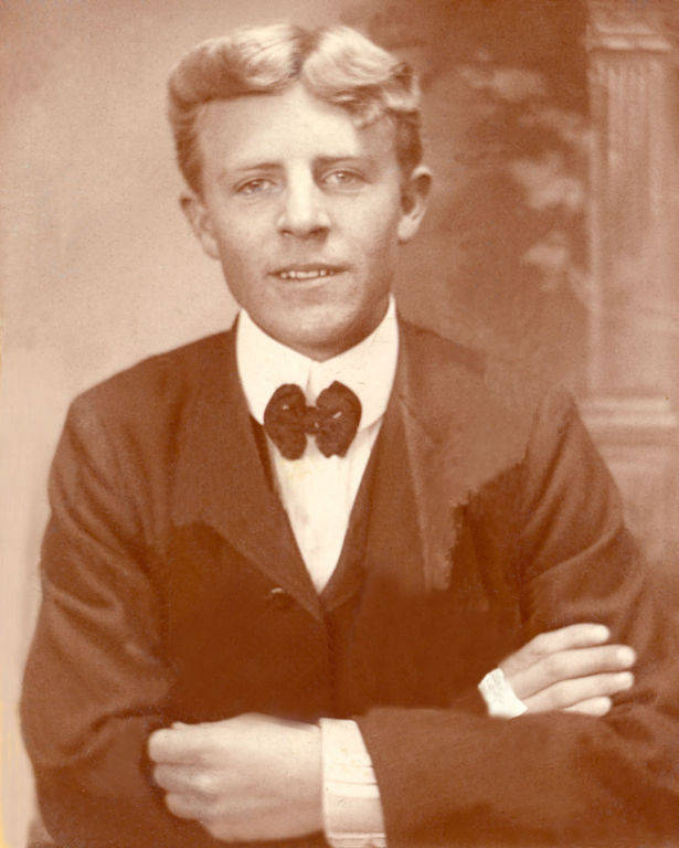 A young man is seated with arms crossed, wearing a suit, vest, and bow tie. His hair is styled with a slight wave, and he looks directly at the viewer with a neutral expression. A decorative column is partially visible in the background.