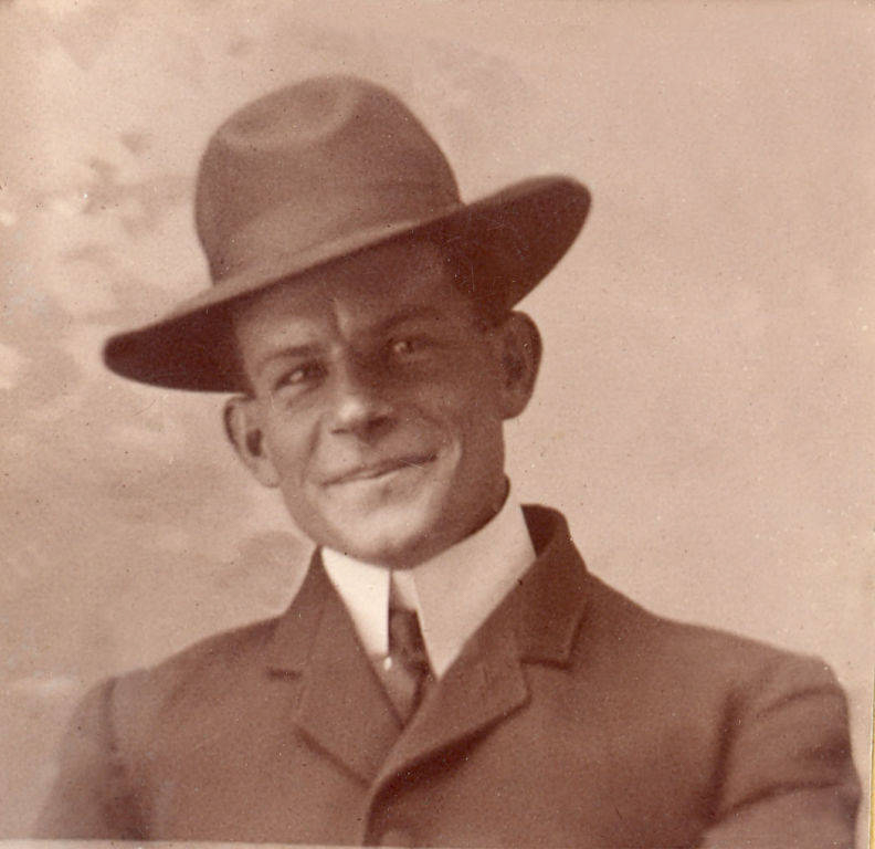 A man wearing a suit, tie, and wide-brimmed hat, smiling slightly. He is turned slightly to the side, with a neutral backdrop behind him.