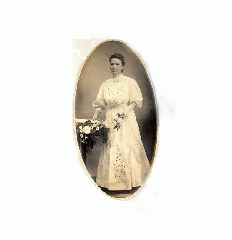 A woman standing and holding a small bouquet of flowers in one hand. She is wearing a long, ornate dress with lace details. Her other hand is resting on a piece of furniture adorned with a larger arrangement of flowers.