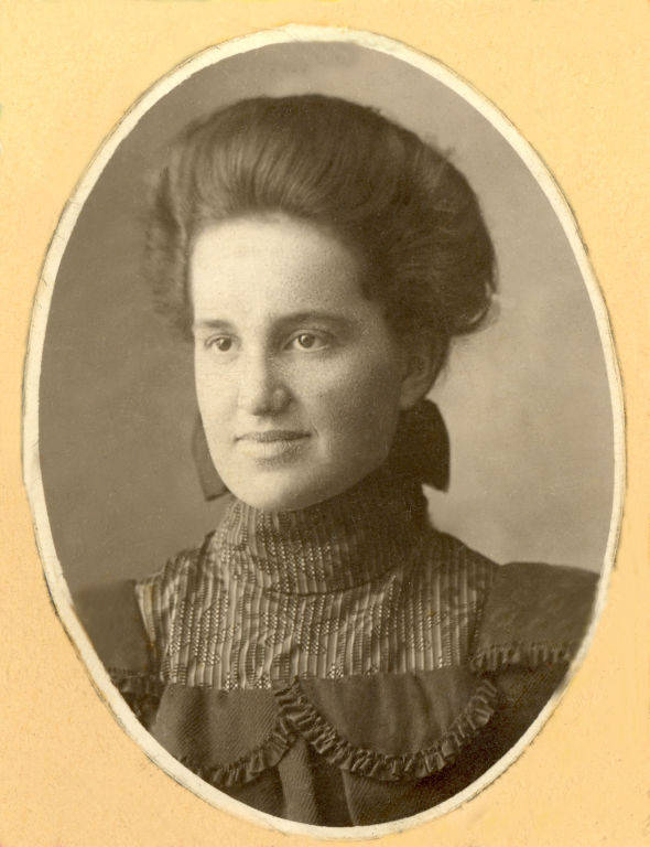 A woman with hair styled in an updo is looking slightly to the side. She is wearing a high-necked dress with a patterned fabric and decorative ruffles. The image is framed in an oval shape. There is no text.