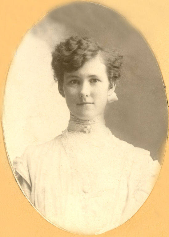 A woman with short, curly hair is wearing a high-necked, ruffled blouse and looking directly at the viewer. The background is plain.