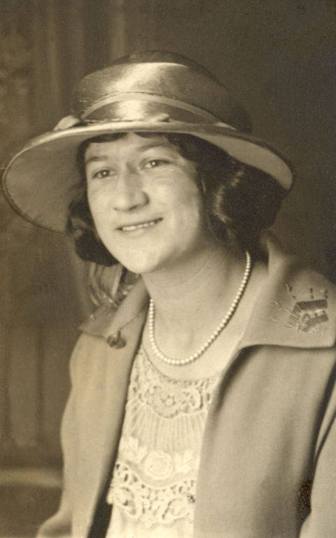 A woman wearing a wide-brimmed hat and a pearl necklace is smiling. She is dressed in a coat with decorative stitching on the collar and a lacy blouse underneath.