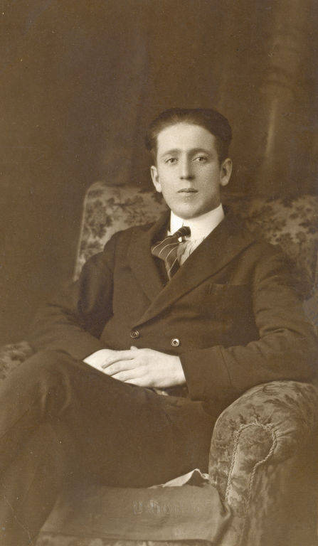 A young man sitting in an ornate, upholstered chair. He is wearing a dark suit with a tie and rests his hands in his lap. The background is simple, with a fabric drape visible.
