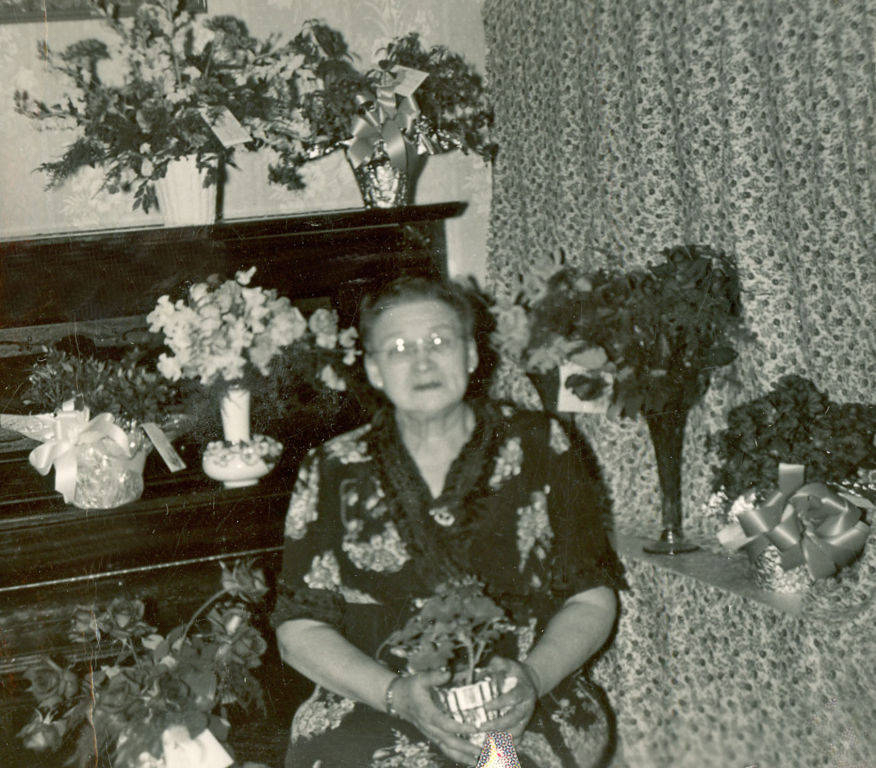 An elderly woman is sitting in a room, holding a small potted plant. She is surrounded by various floral arrangements placed on a piano and a shelf. The room has patterned wallpaper and a curtain. Several of the floral arrangements are decorated with ribbons and cards.