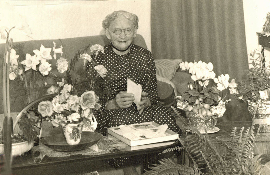 An elderly woman with glasses is sitting on a couch, surrounded by various potted flowers. She is holding a piece of paper and wearing a polka dot dress. There are books and magazines on the table in front of her.