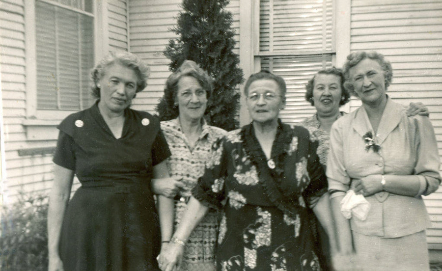 Five women standing together in front of a house with siding. They are wearing dresses and smiling, with some holding hands or linking arms. There is a tree behind them, and windows with shutters are visible on the house.