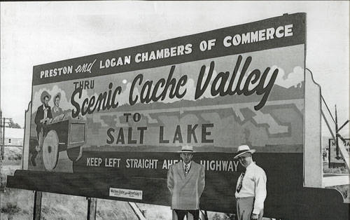 Two men wearing hats and suits stand in front of a large billboard. The billboard reads "PRESTON and LOGAN CHAMBERS OF COMMERCE THRU Scenic Cache Valley TO SALT LAKE KEEP LEFT STRAIGHT AHEAD HIGHWAY." The sign features an illustration of two people and a wagon wheel, with a scenic background.