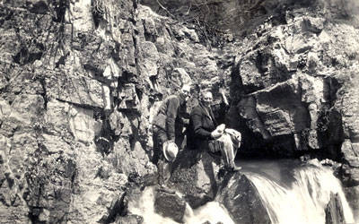 Two people are positioned on a rocky hillside. One person is standing, holding a hat, and the other is sitting near the edge of a small waterfall, with water flowing nearby. The surroundings are rugged and natural.