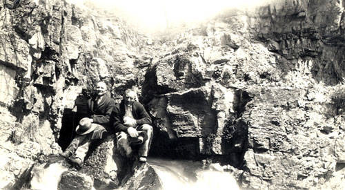 Two people are seated on rocks amidst a rocky cliff or waterfall setting. One person is holding a hat. The scene is framed by large rock formations.