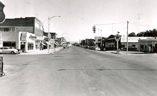 A wide street with shops and businesses on either side, featuring signs and advertisements. Cars are parked along the street. The buildings have signs, including "FURNITURE" and "REXALL DRUGS." There is a Texaco gas station on the right side. Streetlights and utility poles line the area.