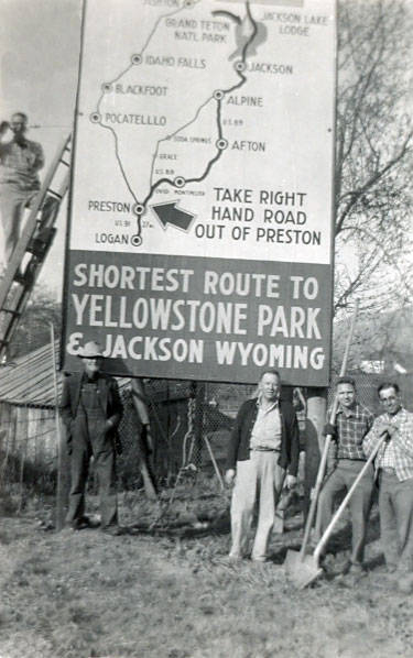 Four men are standing near a large roadside sign, with one man on a ladder. The sign displays a map and reads, "TAKE RIGHT HAND ROAD OUT OF PRESTON." Below, it says, "SHORTEST ROUTE TO YELLOWSTONE PARK & JACKSON WYOMING." One man is holding a shovel and another holds a pickaxe. There is a building and trees in the background.