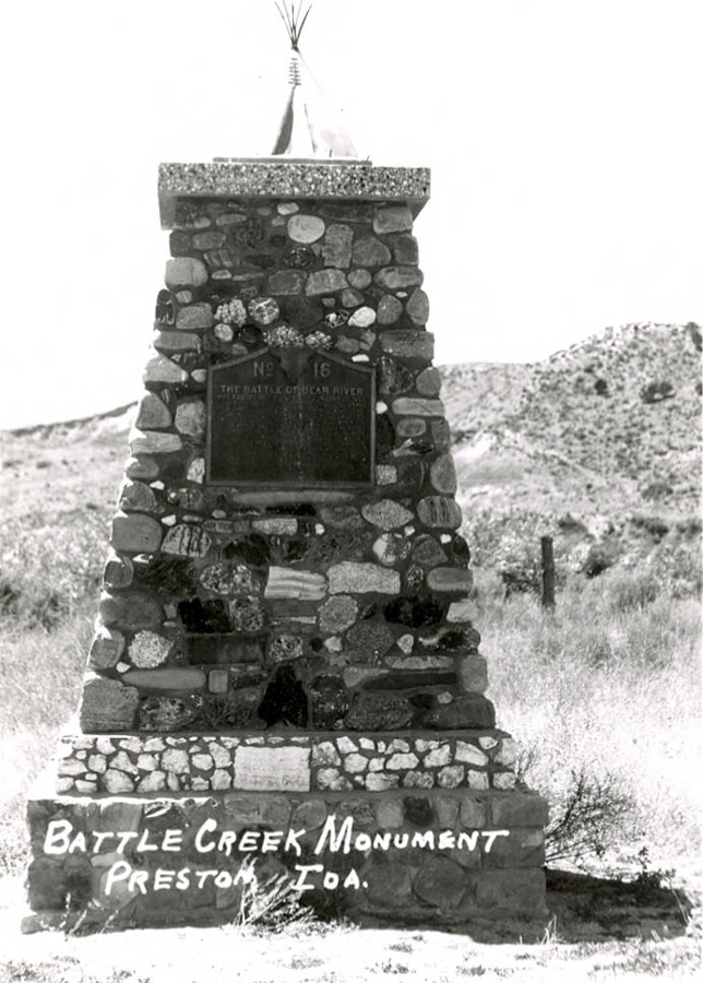 A stone monument in a grassy landscape with hills in the background. The top of the monument is crowned with a small tipi structure. There is a plaque on the front with the text "No. 16 The Battle of Bear River." The base of the monument has the text "Battle Creek Monument Preston, Ida."