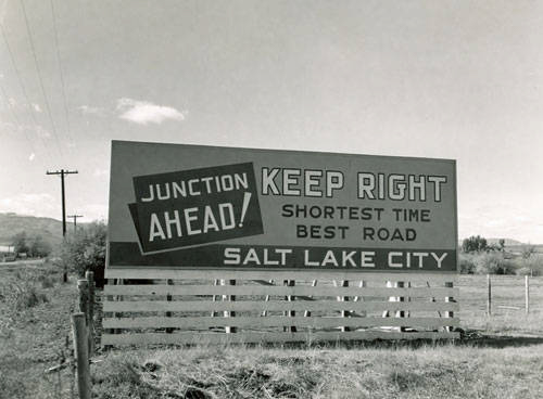 A large roadside sign stands next to a wooden fence in a grassy area with distant hills and utility poles in the background. The sign reads: "Junction Ahead! Keep Right. Shortest Time Best Road Salt Lake City."
