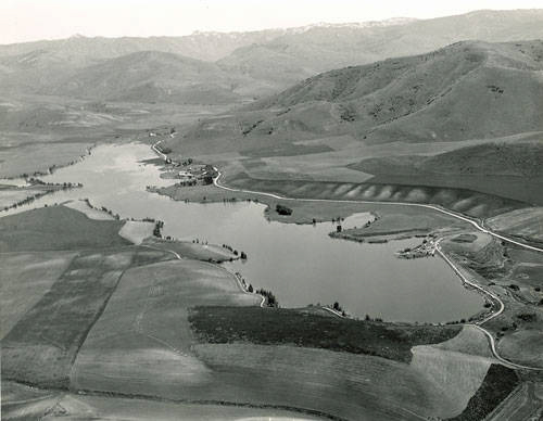 Aerial view of a winding river and a small body of water surrounded by open fields and low hills. The landscape shows a variety of agricultural patterns, with some roads visible near the water. Hills rise in the background, with some covered in patchy vegetation.