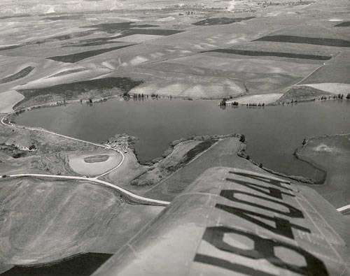 Aerial view of a landscape showing a body of water surrounded by open fields with a few trees. A road curves around the water. Part of an airplane wing is visible in the foreground with the numbers "18404" marked on it.