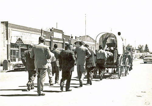 A group of men in suits walk down a street alongside a covered wagon. The street is lined with buildings, and one sign visible reads "Pharmacy." A few cars are parked along the road, and a person is seated inside the wagon. Trees and an open sky are visible in the background.