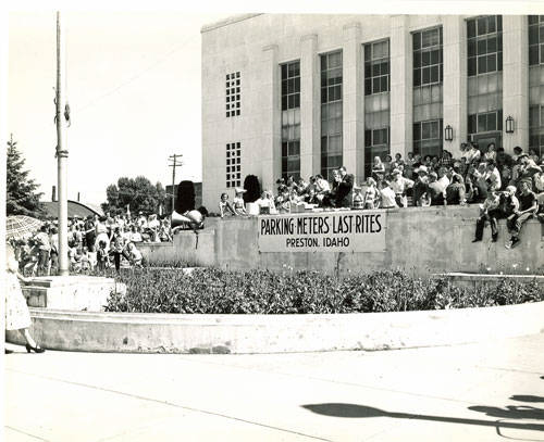 People are gathered in front of a large building with tall windows. Many are seated on a ledge and standing around. There is a banner hanging from the ledge that reads, "PARKING METERS LAST RITES PRESTON, IDAHO." A tall pole is visible on the left, and a landscaped area with bushes is in front of the building.