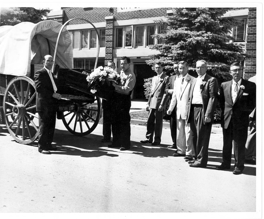 A group of seven men wearing suits stand beside a covered wagon on a street. Two of them hold floral arrangements placed in the wagon. In the background, there is a multi-story brick building with large windows and a tree.
