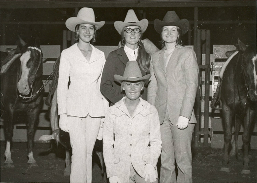 Four women wearing cowboy hats and western attire are posing together. Three are standing, while one is kneeling in front. They are in a stable area with two horses visible on either side. The scene suggests a connection to horse riding or a western-themed event.