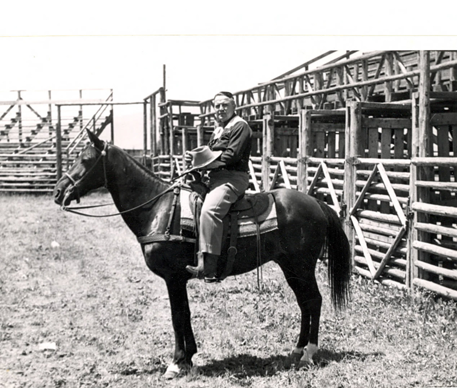A man is sitting on a horse. He is holding a hat in his hand. The setting appears to be an outdoor area with wooden fences and structures in the background.
