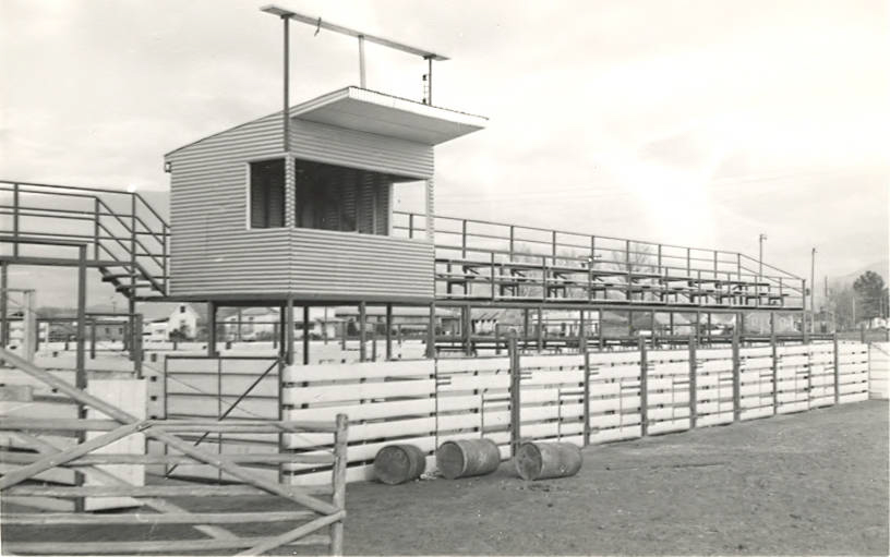 A wooden and metal structure with a small covered booth on top and rows of bleachers, likely a viewing area or announcer's stand. There are several large barrels on the ground nearby. The scene is outdoors, and there is a fenced area surrounding the structure.