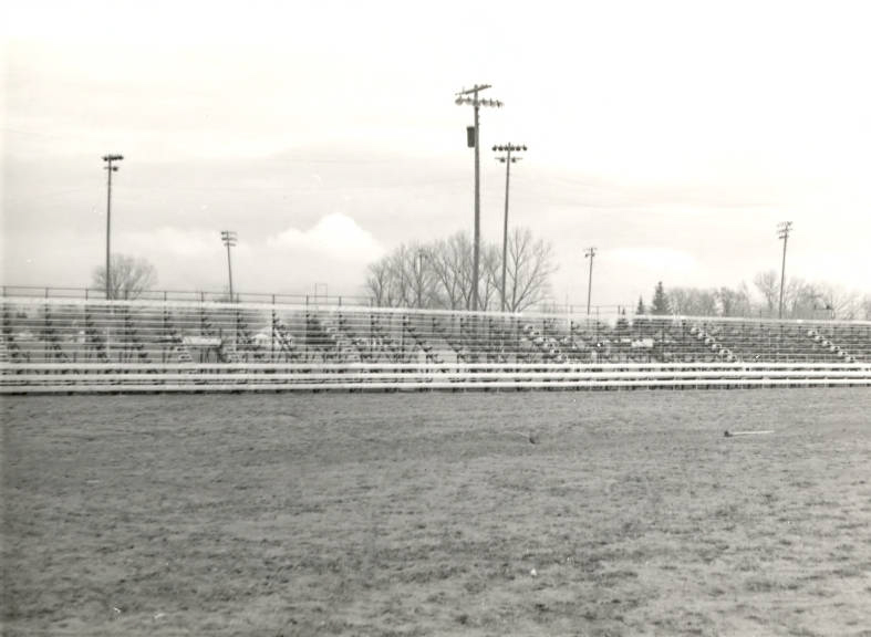 Empty outdoor bleachers in a sports field with several tall light poles and bare trees in the background.