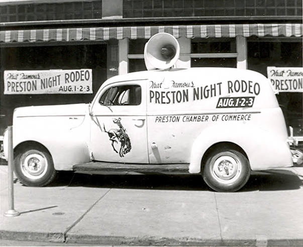 A vintage vehicle is parked on the street. The side of the vehicle displays text reading "That Famous Preston Night Rodeo" with the dates "Aug. 1-2-3." There is an image of a bucking horse and rider on the side. Above the cab is a large speaker. Banners on the building in the background also promote the "Preston Night Rodeo" on the same dates.