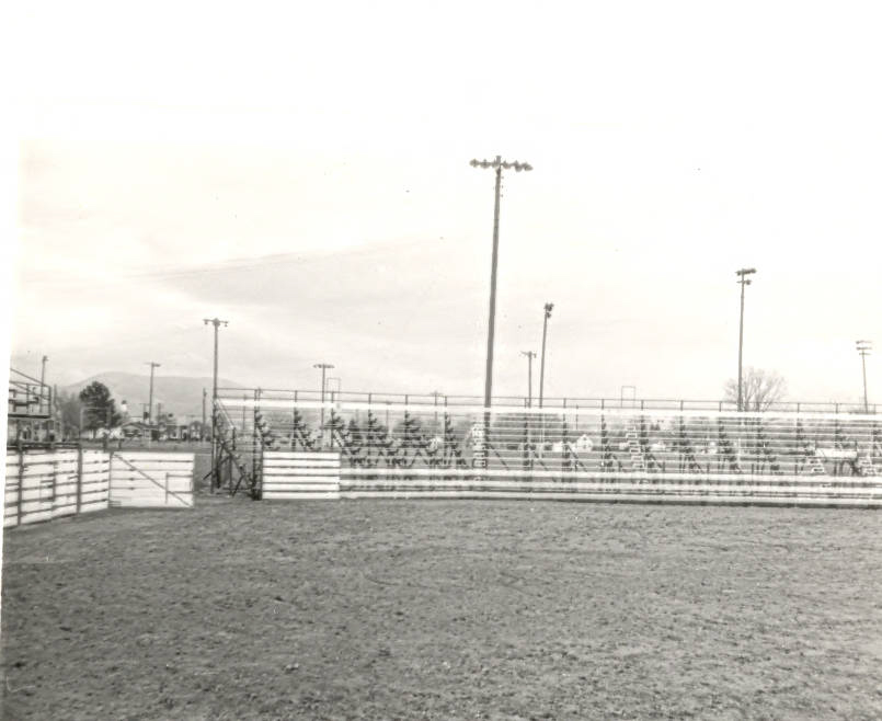 An empty arena with wooden fencing and metal bleachers. Several tall light poles surround the area. In the background, there are trees and some structures visible. The ground is dirt.