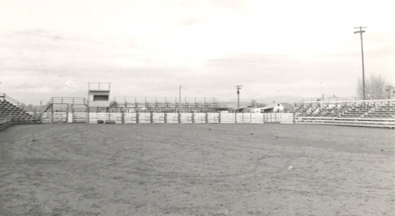 An empty rodeo arena with dirt ground, surrounded by bleachers on three sides. A small booth is visible above the central bleachers. There are some buildings and power lines in the background.
