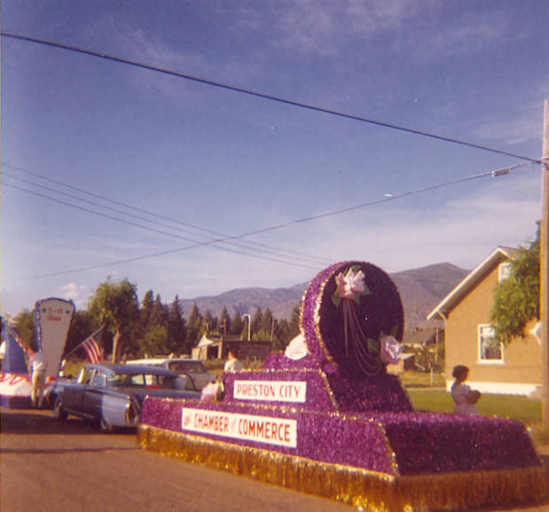 A parade float with purple decorations and white flowers is on a street. The float has signs reading "Preston City" and "Chamber of Commerce." A car with an American flag follows behind. Trees and a house are visible in the background.