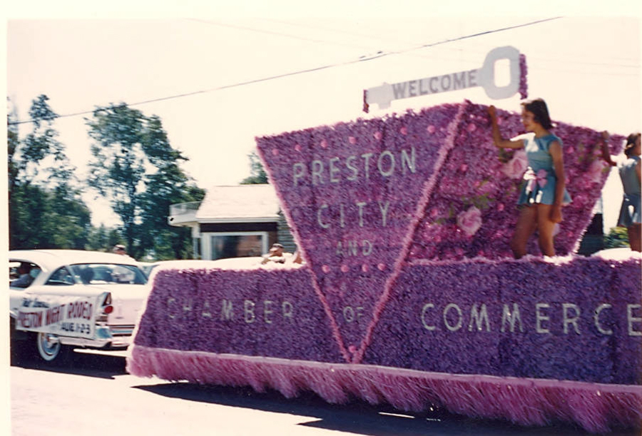 A parade float covered in pink flowers features a large triangular section with the text "PRESTON CITY AND CHAMBER OF COMMERCE." A woman in a blue outfit stands on the float, which is topped with a sign that says "WELCOME." In the background, a car with a sign on the side reads "FRIDAY NIGHT RODEO JULY 1-2-3." A small house and trees are visible in the distance.