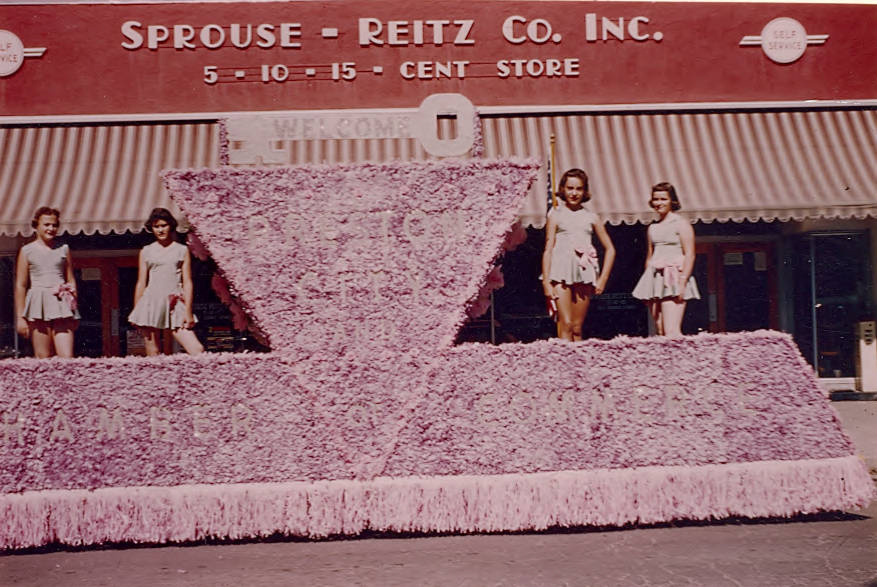 Four women in matching outfits stand on a float decorated with pink flowers. The words "WELCOME," "PRESCOTT," and "CHAMBER OF COMMERCE" are visible on the float. In the background, there is a building with a sign that reads "SPROUSE - REITZ CO. INC. 5 - 10 - 15 - CENT STORE" above an awning.