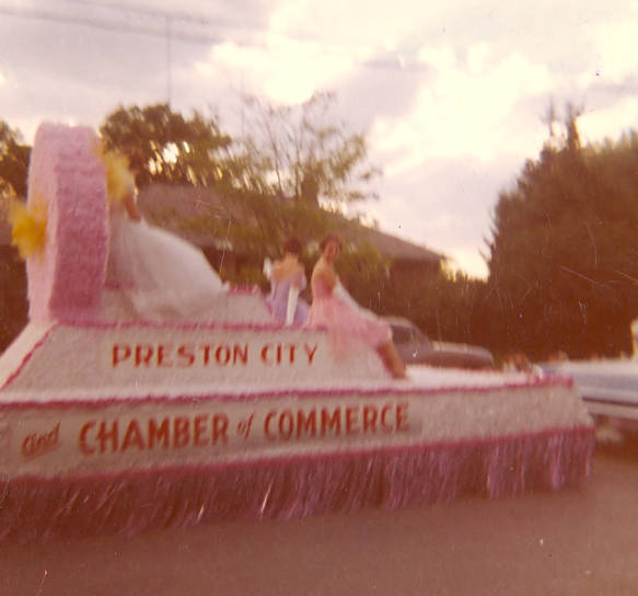 A decorated float with people in formal attire on it, traveling down a street. The float has signage that reads "Preston City and Chamber of Commerce." There are trees and a building in the background.
