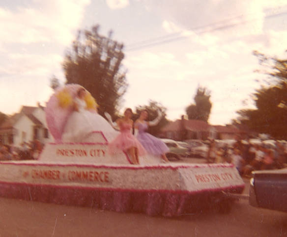 A parade float is moving down a street. It has the words "Preston City Chamber of Commerce" written on it. On the float, women are dressed in colorful dresses, one in pink and another in purple. In the background, there are trees, houses, and a crowd of people watching the parade.