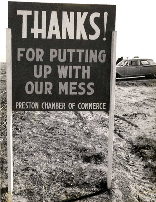 A sign on a dirt road reads, "THANKS! FOR PUTTING UP WITH OUR MESS. PRESTON CHAMBER OF COMMERCE." A car is parked in the background.