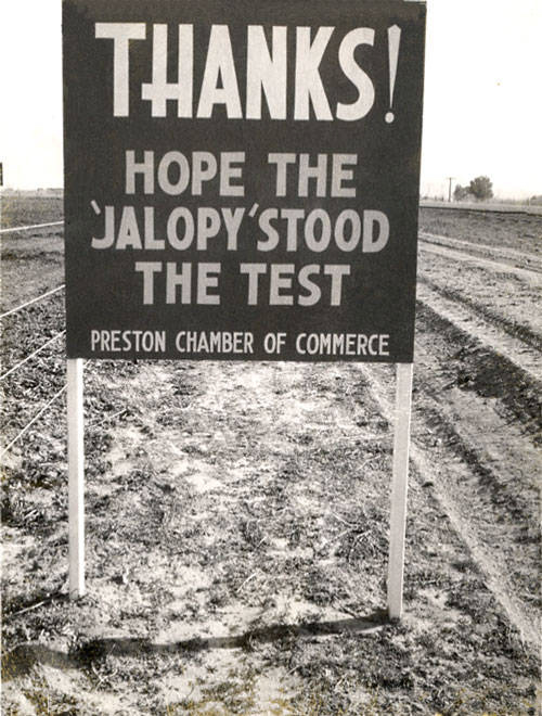 A sign standing on a dirt path reads: "THANKS! HOPE THE 'JALOPY' STOOD THE TEST" with "PRESTON CHAMBER OF COMMERCE" written at the bottom. The background shows open fields and a few distant utility poles.