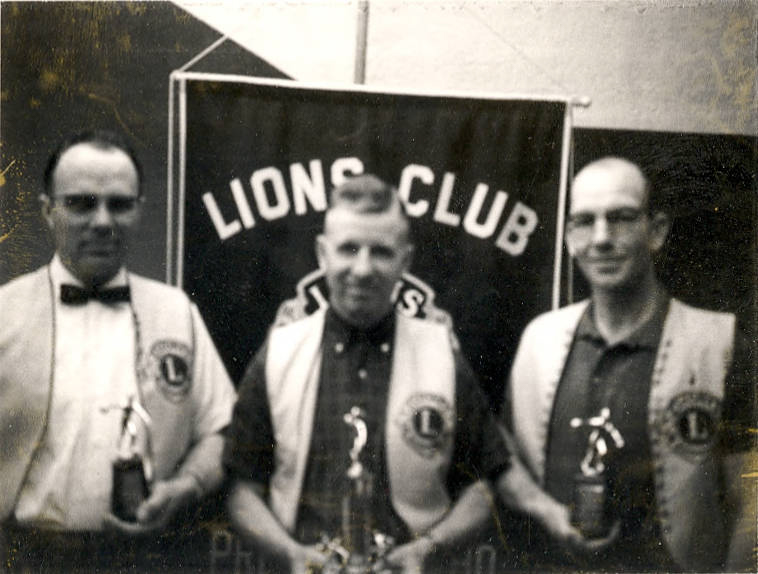 Three men stand holding trophies in front of a "Lions Club" banner. They are wearing vests with an emblem and shirts underneath.