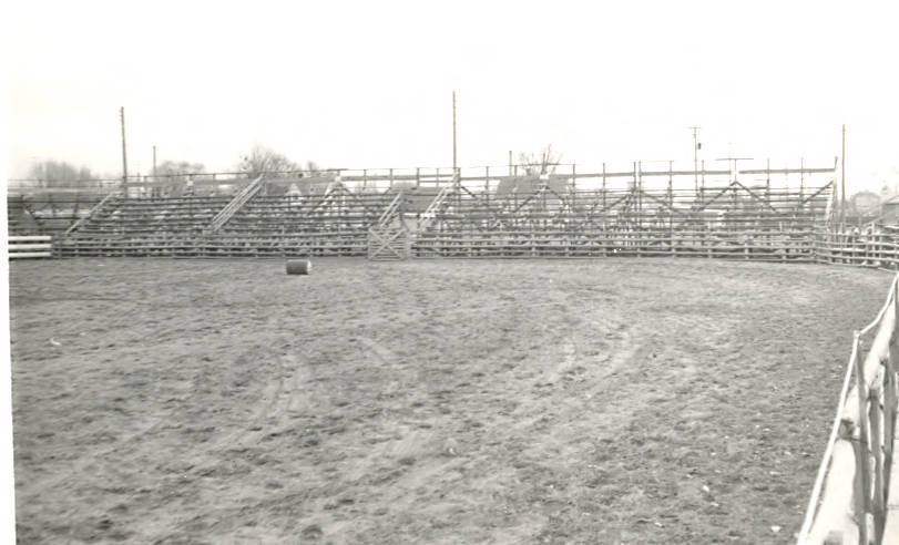 A large, empty arena with wooden bleachers in the background and a flat field in the foreground. A single barrel is placed in the arena. Trees and utility poles are visible beyond the bleachers.
