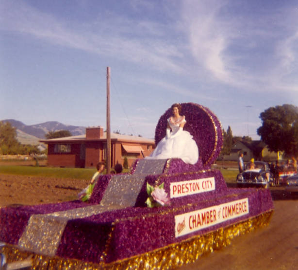 A woman in an elegant gown is seated on a parade float decorated with purple and gold elements. The float displays signs reading "Preston City" and "Chamber of Commerce." There are people and a car visible in the background, along with a red brick building and distant hills.
