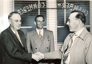 Three men are standing together in front of a window with blinds. The two men on the left and right are shaking hands. The man on the left is wearing glasses and a pinstriped suit. The man on the right is wearing a light-colored coat and striped tie. The man in the middle is wearing glasses and a suit, smiling at the two men shaking hands. Text on the window reads "J. Homan".