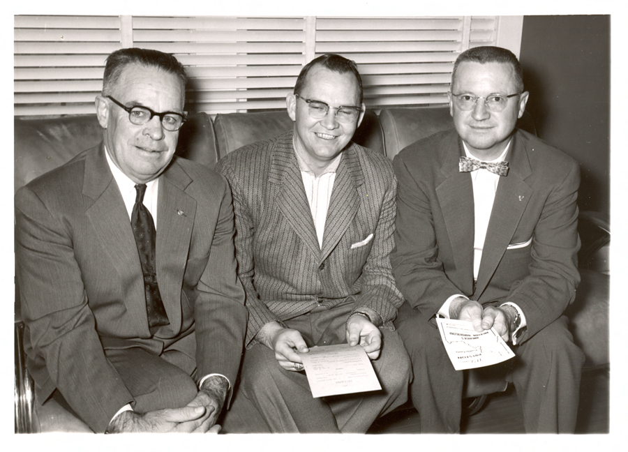 Three men in suits sitting on a couch. The man on the left is wearing glasses and a tie, while the man in the middle is smiling and holding a piece of paper. The man on the right, also wearing glasses, holds a booklet with text partially visible, including "Cheetah" and "Octopus Club." They are seated in front of a window with blinds.