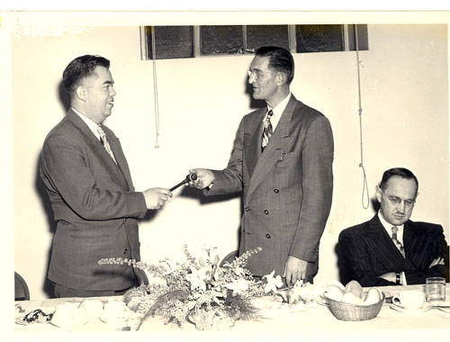 Two men are standing and shaking hands, possibly exchanging an item. They are dressed in suits. A third man is seated at a table with floral arrangements and a bowl of fruit. The setting appears to be a formal event, possibly a meeting or presentation. The seated man is looking down, seemingly uninterested or deep in thought.