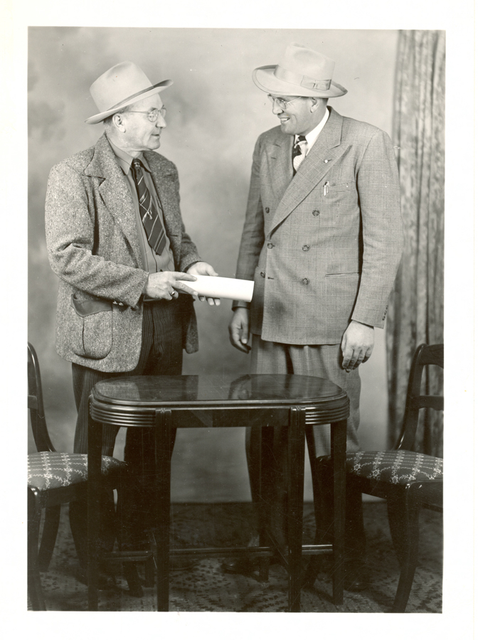 Two men are standing and facing each other next to a small wooden table with patterned fabric chairs on either side. Both are wearing hats and suits. One man, wearing glasses, hands over a rolled-up paper to the other, who is smiling. A curtain is visible in the background.
