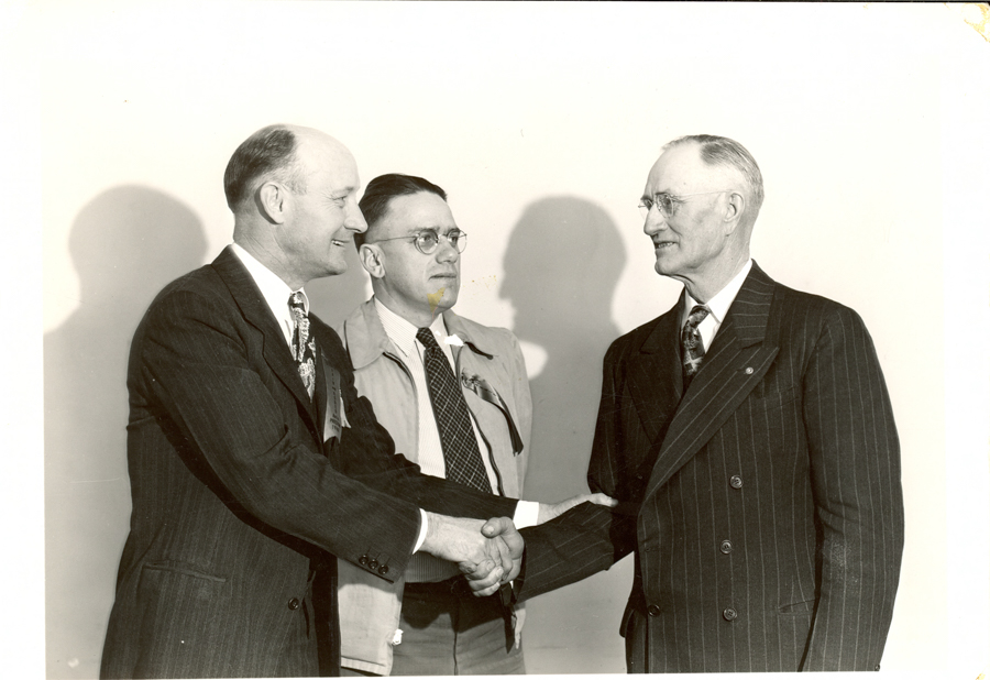 Three men in suits standing together. The man on the left is shaking hands with the man on the right. The man in the middle is wearing glasses and a lighter jacket. They are all smiling and standing in front of a plain background.