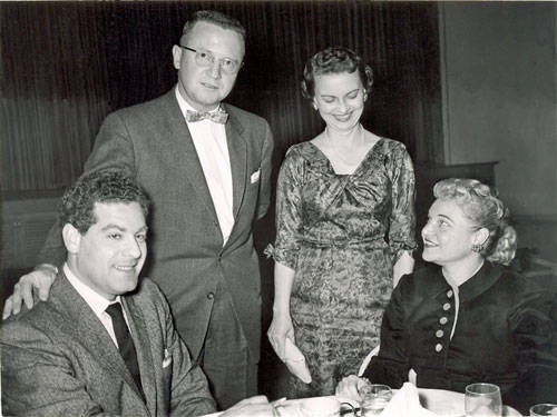 A group of four people are gathered in what appears to be a dining setting. Two men and two women are present. One man is seated at a table next to a woman, both looking in the same direction. The other man stands next to the table, wearing glasses and a bow tie, while the second woman stands beside him, smiling at the seated woman. The table has plates, glasses, and napkins. The background includes a curtain.