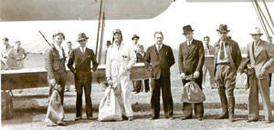 A group of seven men standing in front of an aircraft. One man is wearing a pilot outfit and holding a bag. The others are wearing suits and hats, with some holding bags. In the background, additional people and the aircraft's wings are visible.