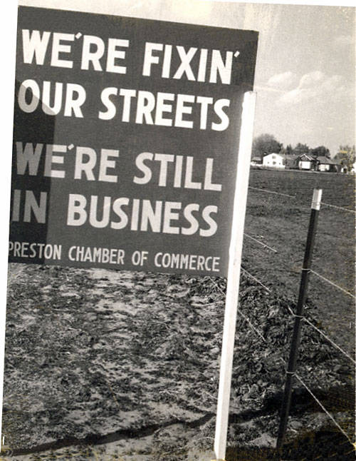 A sign stands beside a dirt path and a wire fence, with open land and houses in the background. The sign reads "WE'RE FIXIN' OUR STREETS WE'RE STILL IN BUSINESS PRESTON CHAMBER OF COMMERCE."
