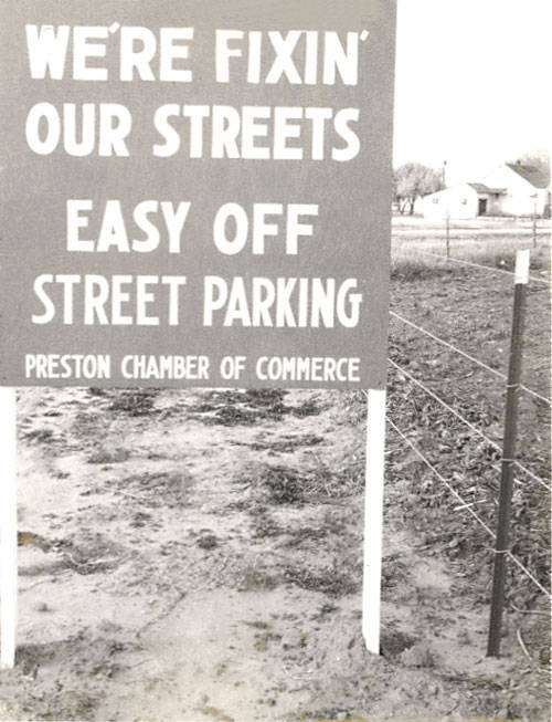 A sign stands near a dirt path and a wire fence, displaying the message: "WE'RE FIXIN' OUR STREETS EASY OFF STREET PARKING" with "PRESTON CHAMBER OF COMMERCE" written below. In the background, there are a few houses and trees.